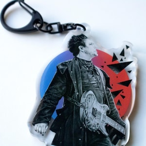 May include: A black and white image of a person holding a guitar, with a red and blue background. The person is wearing a black jacket and a black turtleneck. The guitar is a solid body electric guitar with a black finish. The background is a red and blue circle with a pattern of black triangles.