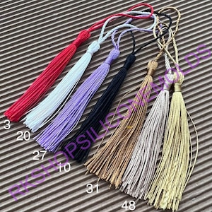 May include: Assortment of eight colorful tassels in red, white, lavender, black, brown, beige, and gold. The tassels are arranged in a row on a brown textured surface.