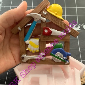 May include: A pink silicone mold shaped like a house with a wooden shed roof. The mold is designed to create a cake or other edible item shaped like a toolbox with tools, including a hammer, saw, wrench, and a circular saw.