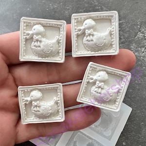 May include: Four white duck-shaped embellishments with a square frame and a bow, along with a silicone mold. The duck design is embossed with details. The items are held in a hand.