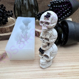 May include: A silicone mold for making a stack of three white skull candle holders. The mold is rectangular and has three cavities shaped like skulls. The mold is next to a stack of three white skull candle holders.