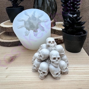 May include: A collection of Halloween-themed items. A white silicone mold in the shape of a spider, a small pile of white skulls, and several black potted plants are arranged on a wooden surface. A dark glass vase is in the background.