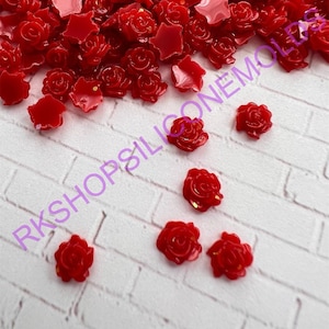 May include: A large quantity of small, red, resin rose embellishments. The roses are scattered on a white brick background. A few roses are also scattered on a white surface.