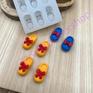 May include: A silicone mold with six cavities shaped like baby shoes. There are six finished resin baby shoes, four yellow with red bows and two blue with red bows.