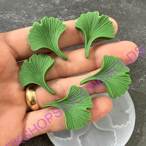May include: Five green ginkgo leaf-shaped resin pieces and a silicone mold. The leaves are a vibrant green with detailed veining. The mold is clear and shows the leaf shape. The items are held in a hand.
