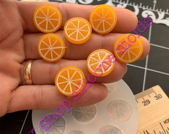 Puede incluir: Un molde de silicona con seis cavidades con forma de rodajas de naranja. El molde es transparente y las cavidades están llenas de resina de color naranja. También hay varias piezas de resina con forma de rodajas de naranja junto al molde.