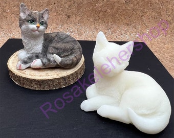 May include: Two ceramic cat figurines, one gray and white and one white. The gray and white cat is sitting on a wooden base. The white cat is sitting on a black surface.