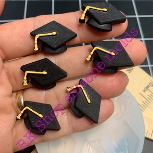 May include: A silicone mold with five impressions of graduation caps. The caps are black with gold tassels. The mold is on a white surface.
