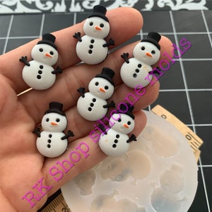 May include: A clear silicone mold with six cavities shaped like snowmen.  There are six white snowman figurines with black hats and black eyes, noses, and buttons.  The mold is on a wooden ruler with inch markings.