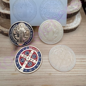 May include: An assortment of Saint Benedict medals and a mold. The medals are in various colors, including gold, red, blue, and white. The mold is clear and contains two circular impressions of the medal design. The medals feature religious text and imagery.