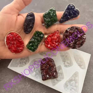 May include: A set of nine silicone molds for making resin jewelry. The molds are shaped like different geometric shapes, including a heart, a diamond, a circle, and a teardrop. The molds are shown with finished resin pieces in various colors, including red, green, blue, and purple.