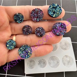 May include: A close-up of a hand holding seven iridescent agate cabochons, with a silicone mold for making cabochons in the background. The cabochons are various shades of blue, purple, and green.