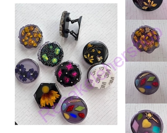 May include: Assortment of 10 phone grips with various designs. The grips are round, hexagonal, and square. Some designs include glitter, pressed flowers, and colorful geometric shapes. The grips are made of clear resin and have a black adhesive backing.