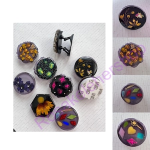 May include: Assortment of 10 phone grips with various designs. The grips are round, hexagonal, and square. Some designs include glitter, pressed flowers, and colorful geometric shapes. The grips are made of clear resin and have a black adhesive backing.