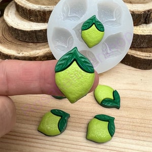 May include: A silicone mold with five cavities shaped like lemons with leaves. There are four green lemons with leaves on a wooden surface.