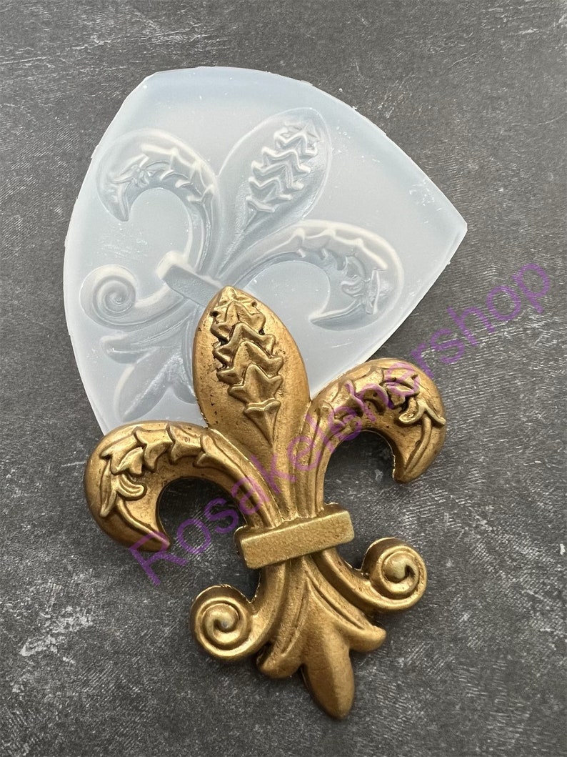 May include: A gold-colored metal fleur-de-lis design with intricate details. The design is shown next to a clear silicone mold of the same design.