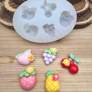 May include: A silicone mold with fruit shapes, including a strawberry, pineapple, grapes, and cherries. The image also shows finished resin fruit charms in pink, purple, red, and yellow, each with a flower accent. The charms are on a wooden surface.