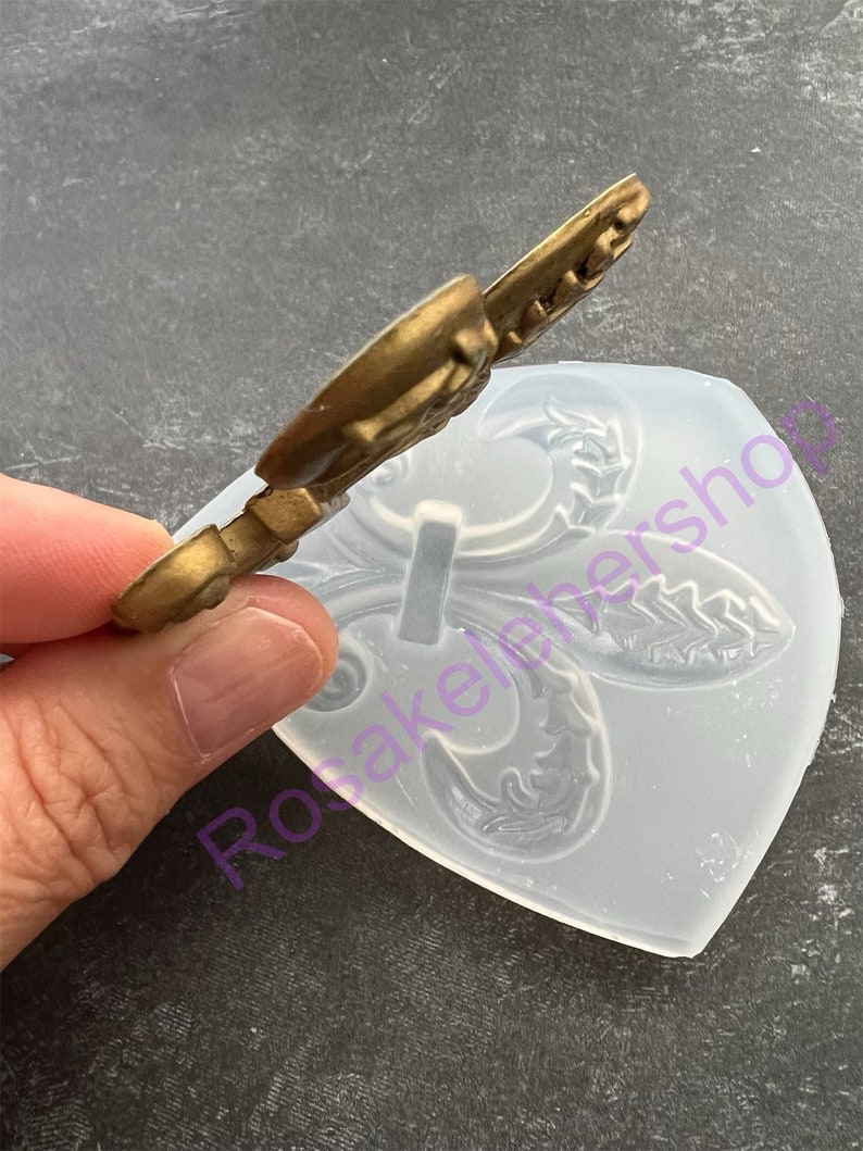 May include: A clear silicone mold with a raised design of a fleur-de-lis. The mold is used for making decorative items like cake toppers or fondant decorations.