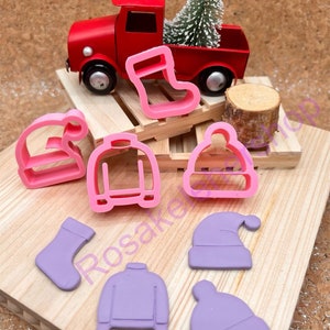 May include: A red toy truck carries a small Christmas tree. Pink and purple cookie cutters in the shapes of a stocking, sweater, and hats are arranged on a wooden surface. A small wooden log is also present.
