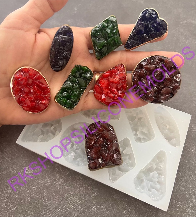 May include: A set of nine silicone molds for making resin jewelry. The molds are shaped like different geometric shapes, including a heart, a diamond, a circle, and a teardrop. The molds are shown with finished resin pieces in various colors, including red, green, blue, and purple.