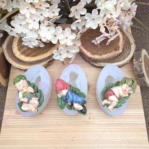 May include: Three silicone molds featuring sleeping baby gnome figurines. Each gnome is resting on a green leaf. One gnome wears a red hat and blue outfit, while the others have green hair and are in various poses. The molds are on a wooden surface.