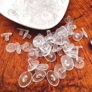 May include: A collection of clear, plastic earring backs, also known as earring stoppers, are scattered on a wooden surface and in a white dish. The earring backs have a disc shape with a post that inserts into the earring.