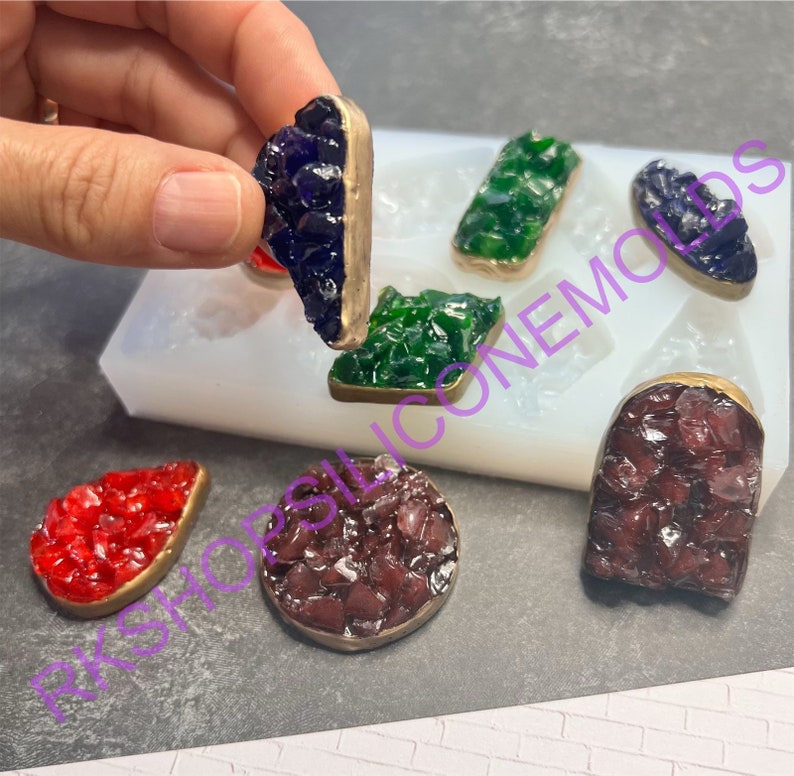 May include: Six resin crystal shapes in various colors, including red, green, purple, and brown, are displayed on a white silicone mold. Each crystal shape has a gold-colored metal rim.