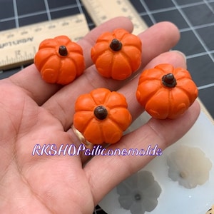 May include: Four small, orange pumpkin-shaped silicone molds with dark brown stems. The molds are held in a hand, with a silicone mold tray and ruler in the background. The text "RKSHOPsiliconemolds" is visible.