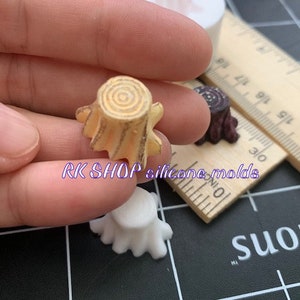 May include: A close-up of a hand holding a small, detailed, light brown tree stump mold. The mold is made of silicone and is designed for crafting. The mold is sitting on a black and white checkered surface.