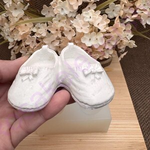 May include: A pair of white baby shoes with a bow detail. The shoes are made of a soft, pliable material and are perfect for a newborn baby.