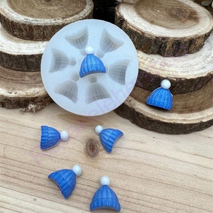 May include: A silicone mold with six cavities shaped like blue knit hats with white pom-poms. There are five blue knit hats with white pom-poms on a wooden surface.