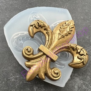 May include: A gold-colored fleur-de-lis design in a clear silicone mold. The mold is shaped like a shield.