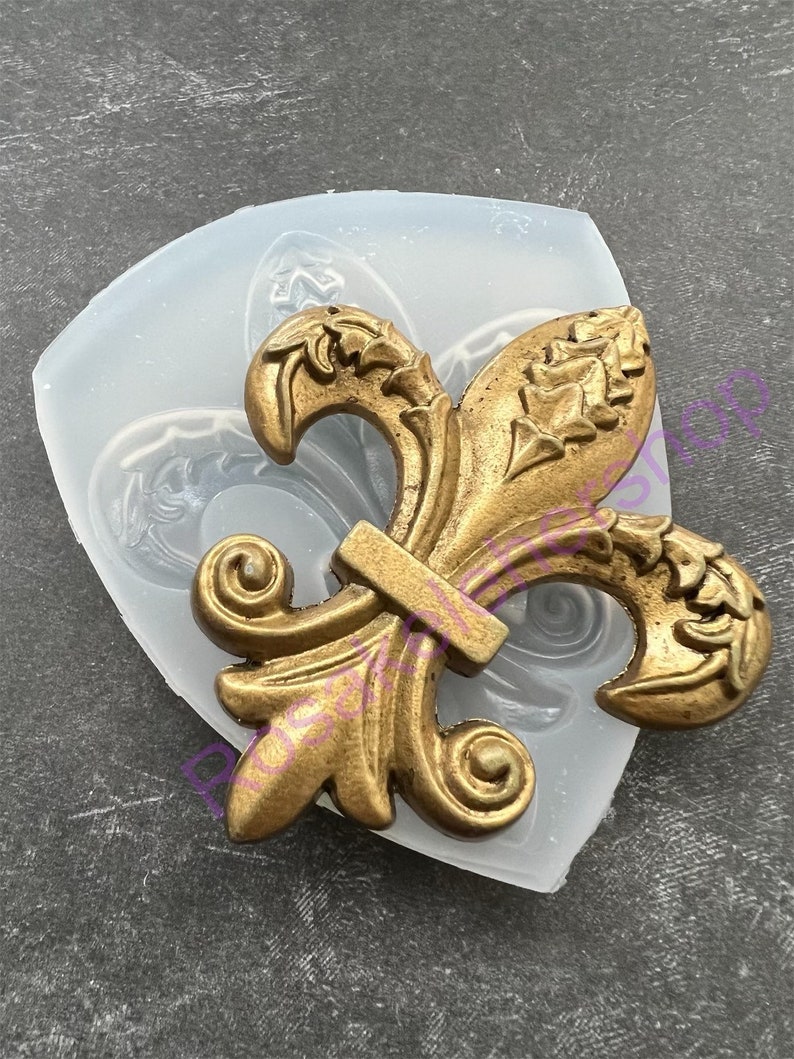 May include: A gold-colored fleur-de-lis design in a clear silicone mold. The mold is shaped like a shield.