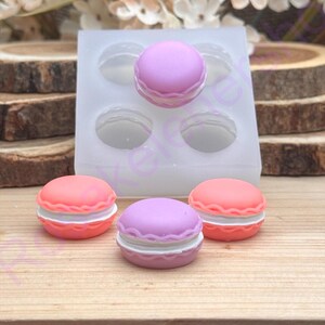 May include: A silicone mold with four cavities, each shaped like a macaron. Three macarons are visible: two in coral and one in lavender, each with a white filling. The macarons sit on a wooden surface.