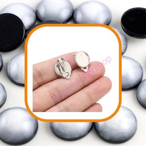 May include: Two silver-toned earring clips with a round, clear, glass-like surface. The clips are held in a hand.