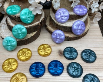 May include: A collection of moon-faced cabochons in various colors, including teal, gold, blue, and purple. The cabochons are arranged on a wooden surface, with some placed on small wooden slices. White floral accents are in the background.