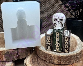 Puede incluir: Una figurita de calavera blanca con una pila de libros verdes. Los libros están etiquetados con "Bats", "Brews" y "Book". La calavera está sentada sobre una base de madera. Hay un molde de silicona en el fondo.