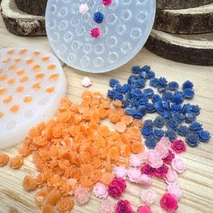 May include: A collection of colorful, miniature rose-shaped embellishments in shades of orange, blue, pink, and clear. The image also shows two circular molds with rose-shaped cavities, one filled with orange roses and the other with a few pink and blue roses.