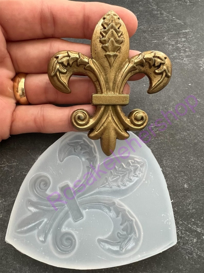 May include: A gold-colored metal fleur-de-lis design with intricate details. The design is shown next to a clear silicone mold of the same design.