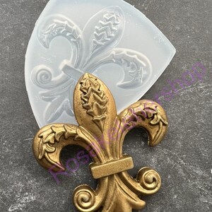 May include: A gold-colored metal fleur-de-lis design with intricate details. The design is shown next to a clear silicone mold.