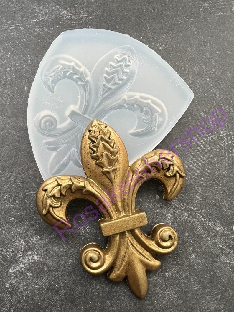 May include: A gold-colored metal fleur-de-lis design with intricate details. The design is shown next to a clear silicone mold.