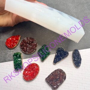 May include: A clear silicone mold for making resin jewelry. The mold is shown with several finished pieces of jewelry, including a red, green, and blue heart-shaped piece, a round piece, and a rectangular piece.
