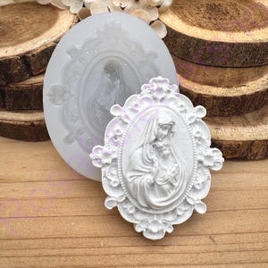 May include: A white, oval-shaped silicone mold and a finished white resin cameo. The cameo features a religious figure holding a child, framed by an ornate floral border. The mold is round with a similar design.
