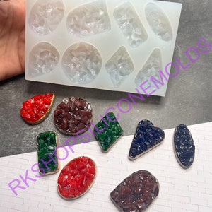 May include: A silicone mold with eight different shapes for making resin jewelry. The mold is white and has a textured surface. The shapes include a circle, a square, a rectangle, a triangle, a heart, a teardrop, a diamond, and a star. The mold is shown with several finished resin pieces in different colors.
