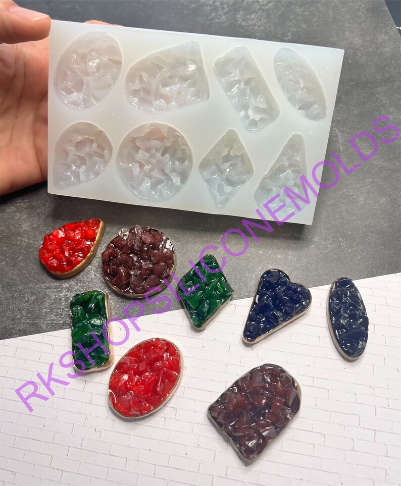 May include: A silicone mold with eight different shapes for making resin jewelry. The mold is white and has a textured surface. The shapes include a circle, a square, a rectangle, a triangle, a heart, a teardrop, a diamond, and a star. The mold is shown with several finished resin pieces in different colors.