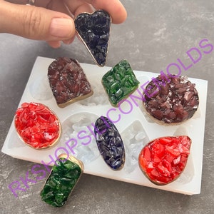 May include: Silicone mold with eight different shapes for making resin jewelry. The mold is white and has a variety of shapes, including a heart, a rectangle, an oval, and a teardrop. The mold is filled with colorful resin pieces.