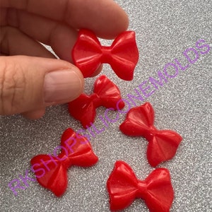 May include: Five small, glossy red bow-shaped embellishments are scattered on a glittery silver surface. One bow is held by a hand. The bows are evenly colored and appear to be made of a hard plastic or resin material.