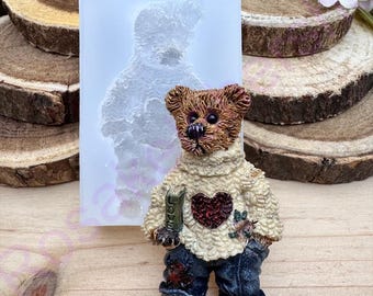 May include: A brown teddy bear wearing a white sweater with a red heart and blue jeans. The bear is holding a book that says "Love" on the cover. The bear is standing on a wooden surface.
