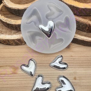 May include: A clear silicone mold with heart-shaped cavities, containing a silver heart. Four additional silver hearts are scattered around the mold. The items are on a wooden surface, suggesting a craft or jewelry-making theme.