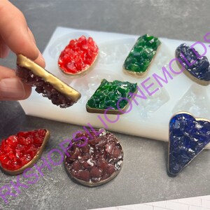 May include: Silicone mold with six different shapes for making resin jewelry. The mold is filled with red, green, blue, and brown resin pieces. The resin pieces are shaped like a heart, a rectangle, a diamond, a circle, and two teardrop shapes.
